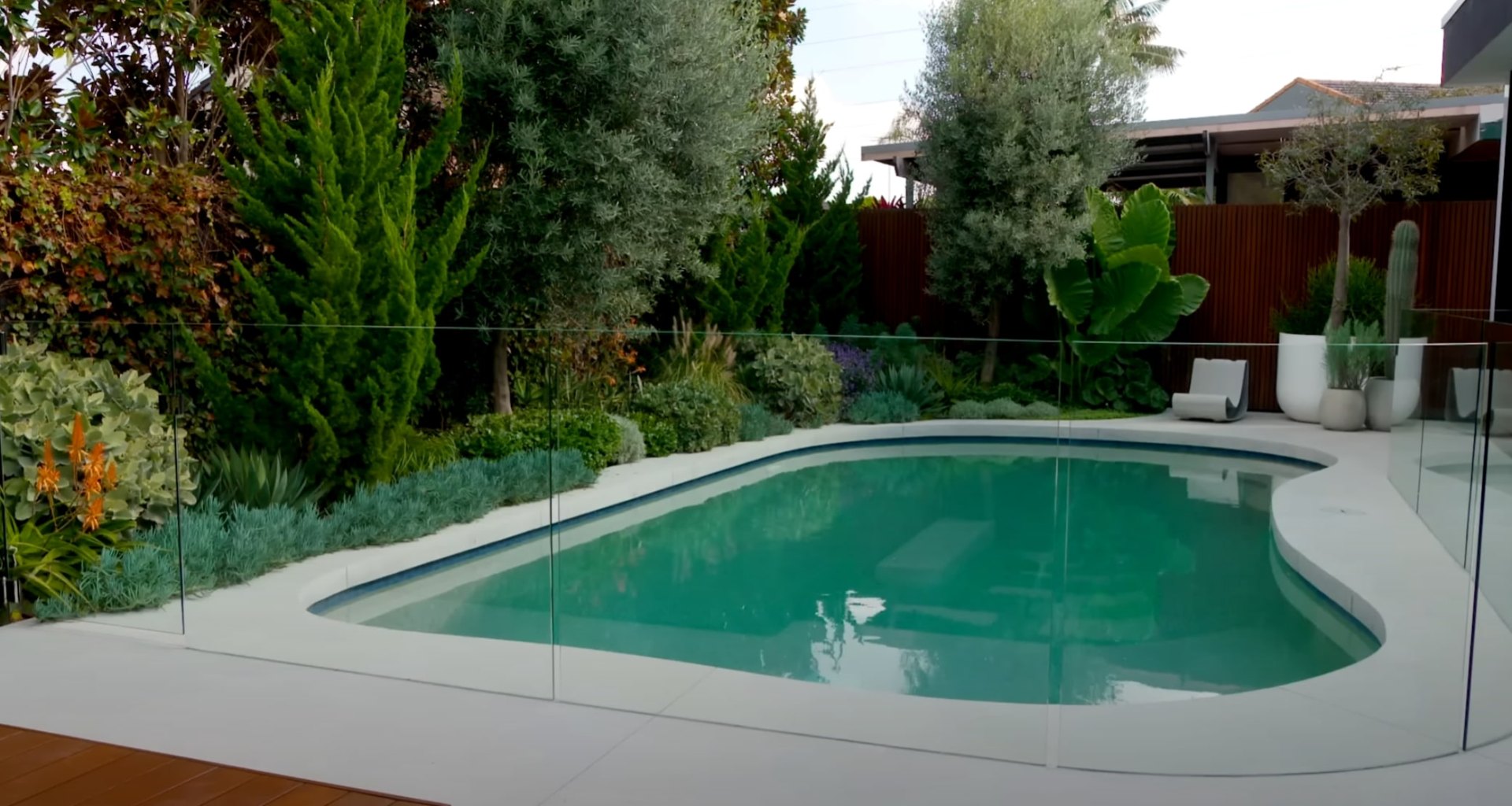 Pool Area Landscaping with Glass Fencing and Tropical Plants Modern backyard with swimming pool, glass safety fencing, and professional landscaping featuring diverse plants and clean hardscaping